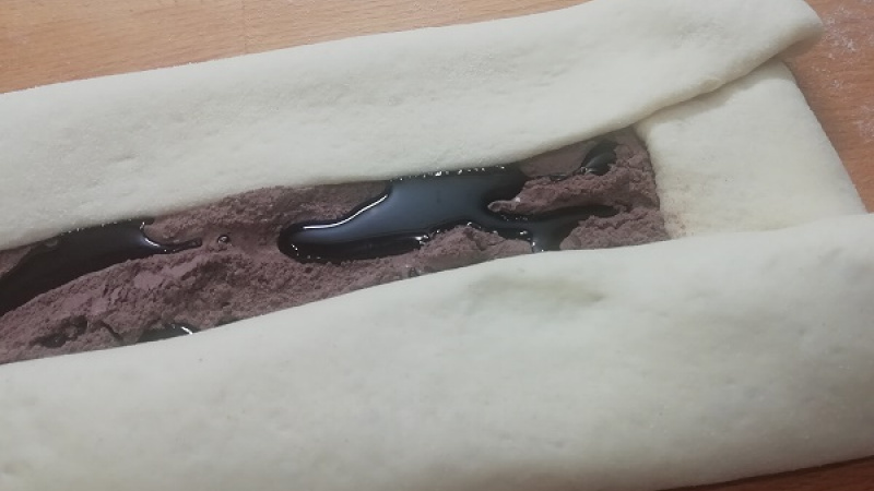 4. Kakao zmiešame s práškovým cukrom. Maslo roztopíme. Cesto rozvaľkáme na 1 cm hrúbku, veľkosť cca 45x25 cm. Cesto potrieme maslom (nešetríme). Okraje cca 5 cm vynecháme. Na maslo posypeme polovicu kakaovej náplne, opäť pokvapkáme maslom.